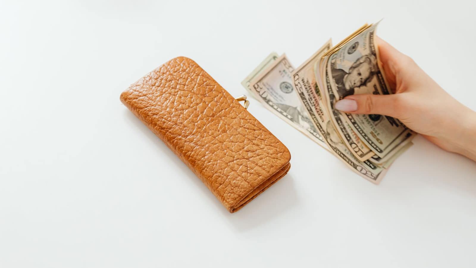 A hand counting US dollar bills beside a leather wallet on a pale surface